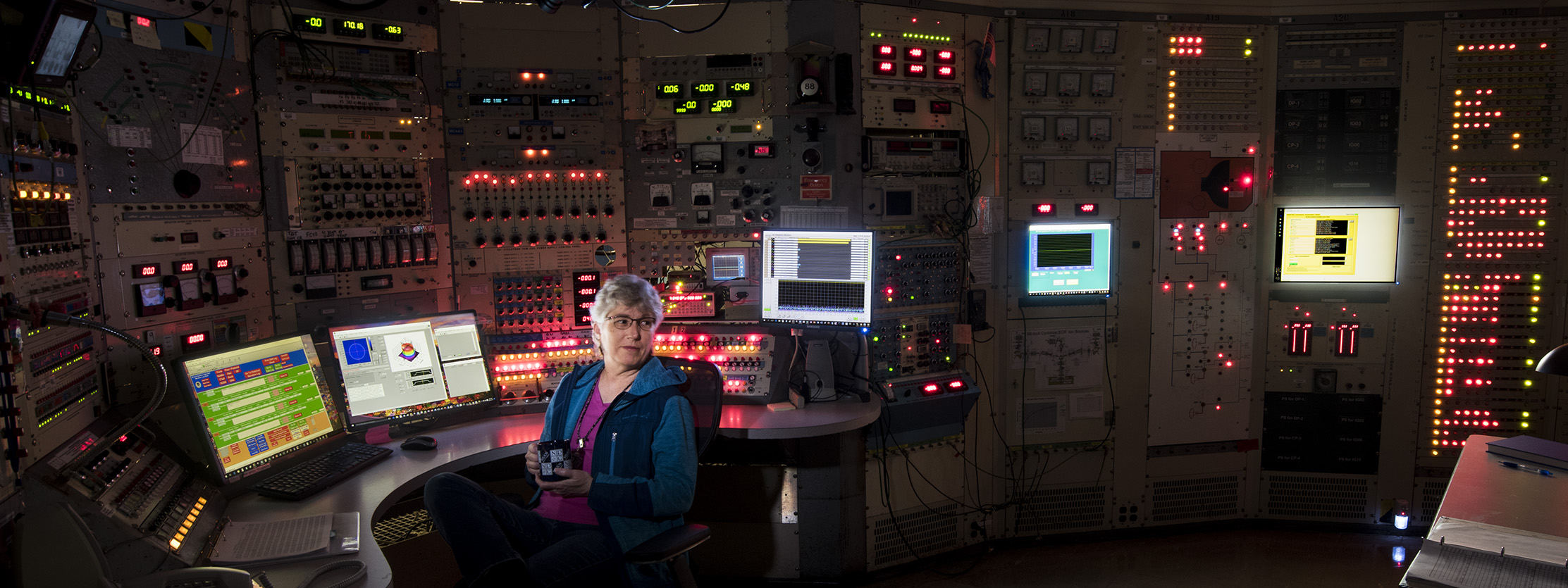 Operator of the 88-Inch Cyclotron sits in a dark control room surrounded by computer screens and glowing panels of lights