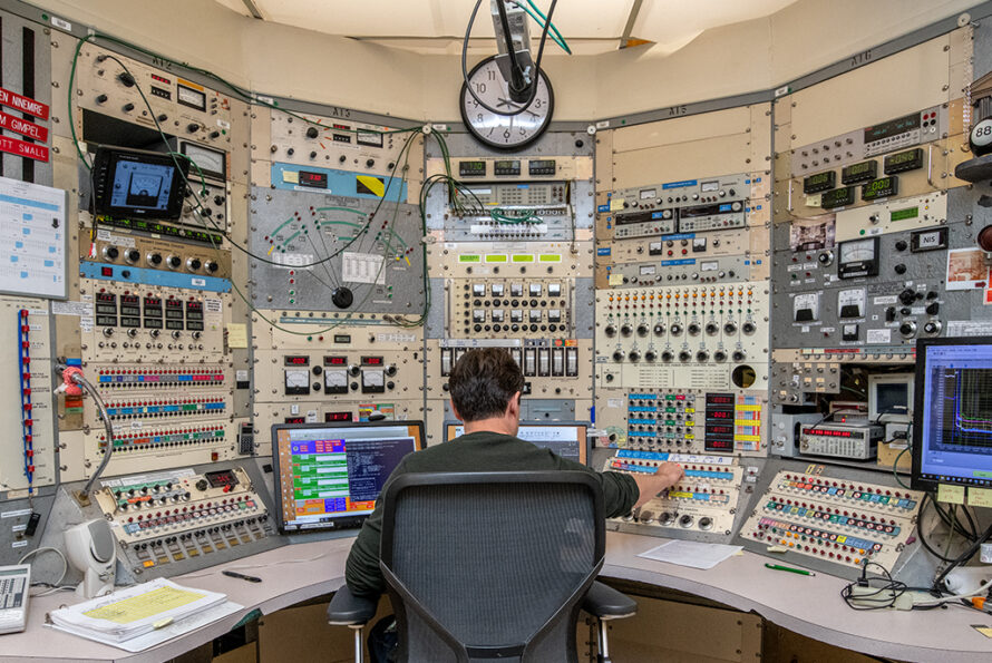 A person adjusts a setting at a console with multiple panels of knobs, switches, and screens.