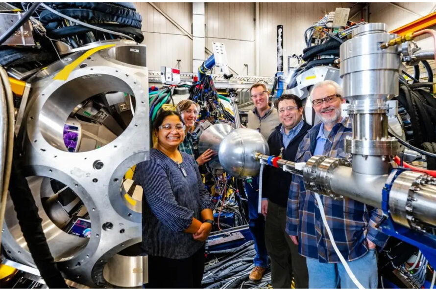 A group of five people (two women and three men) stand next to a large, spherical scientific machine.