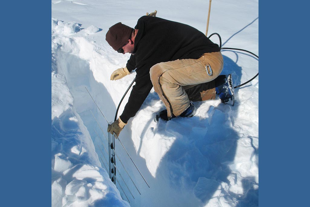 A white male in a black top and beige pants lowers equipment into a hole in the snow and ice.