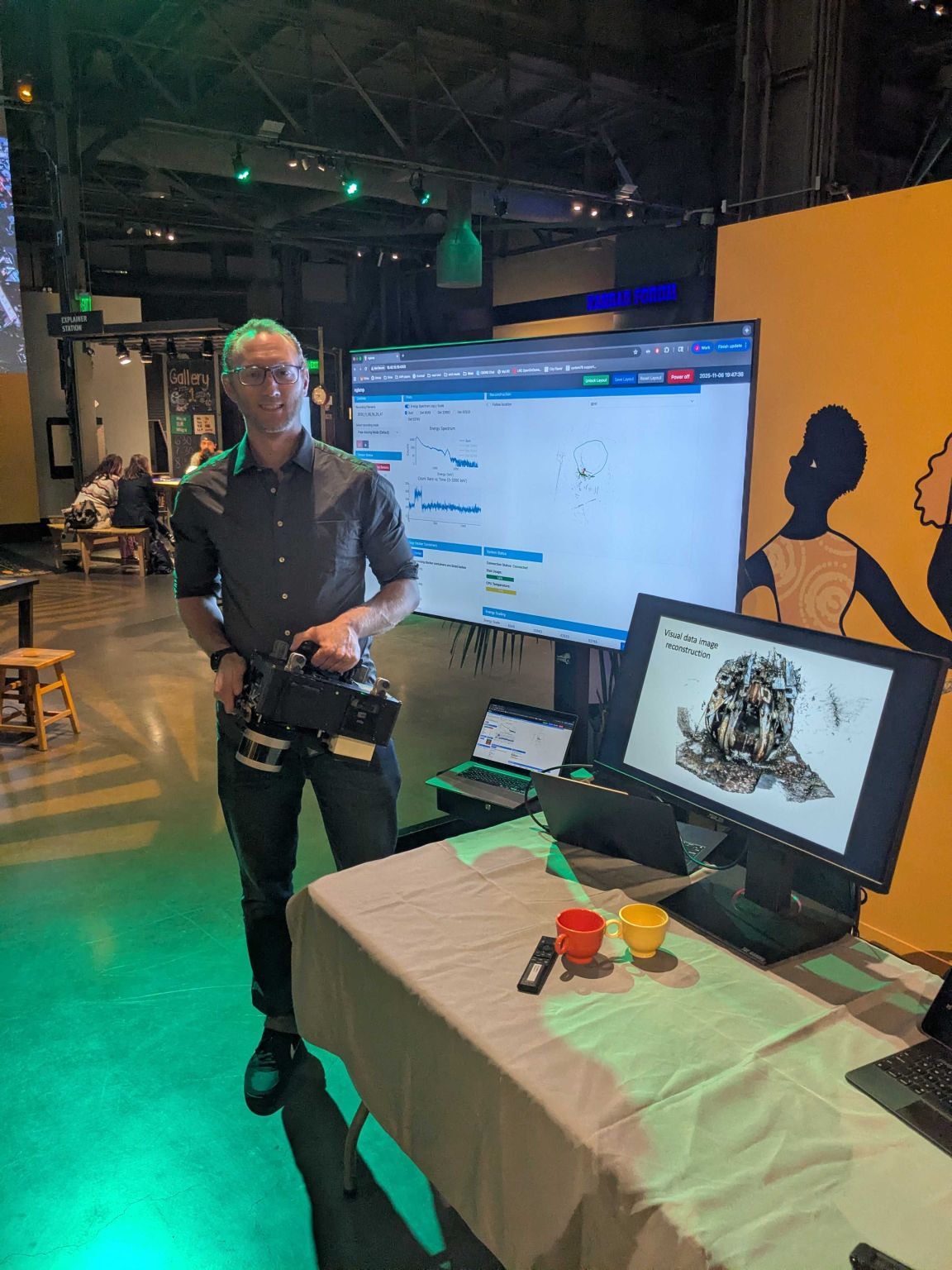 A while male in grey dress shirt holding a piece of equipment in front of displays showing scientific plots and imagery.