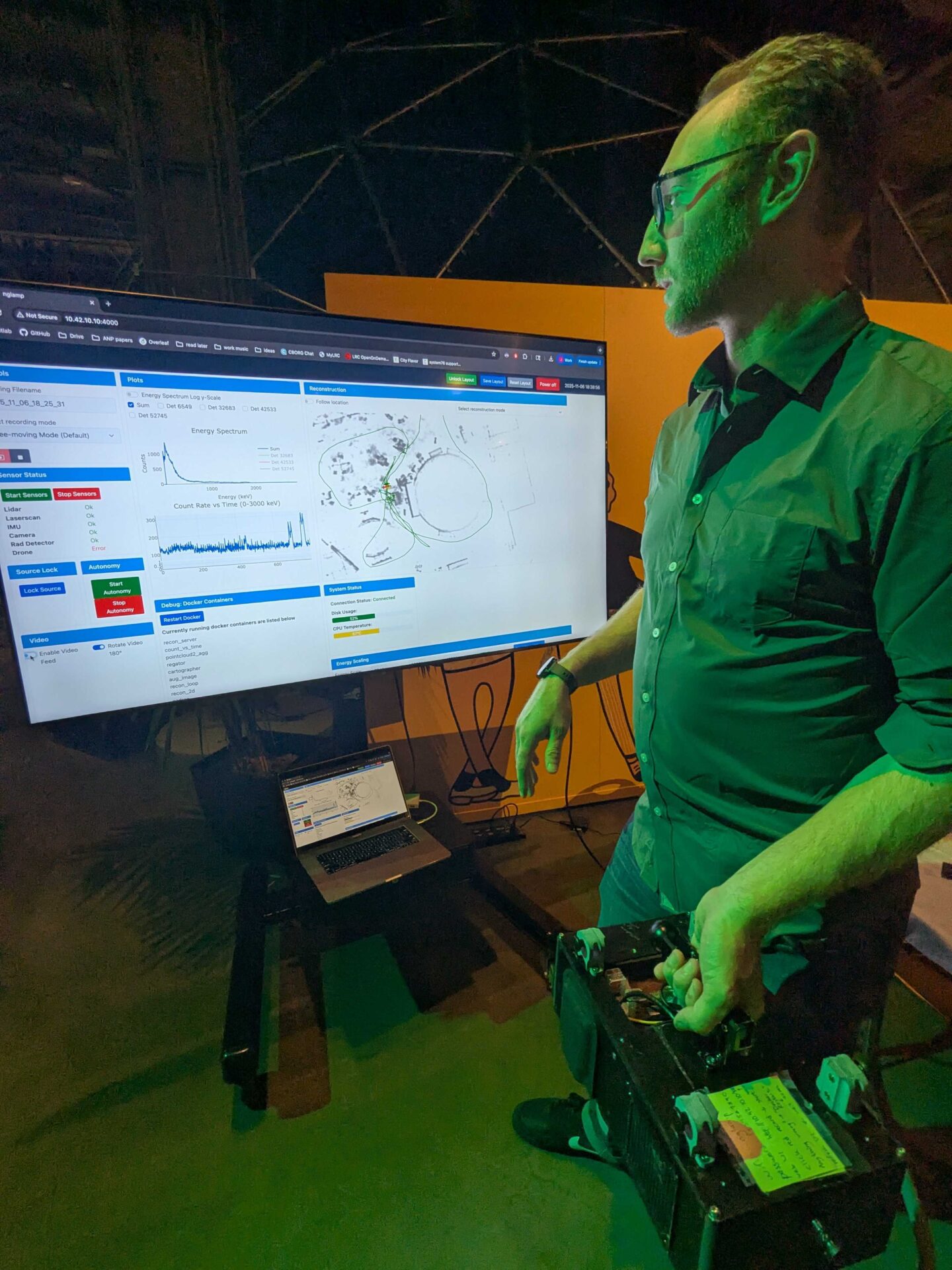 A white male holding an electronic device in front of a digital display.
