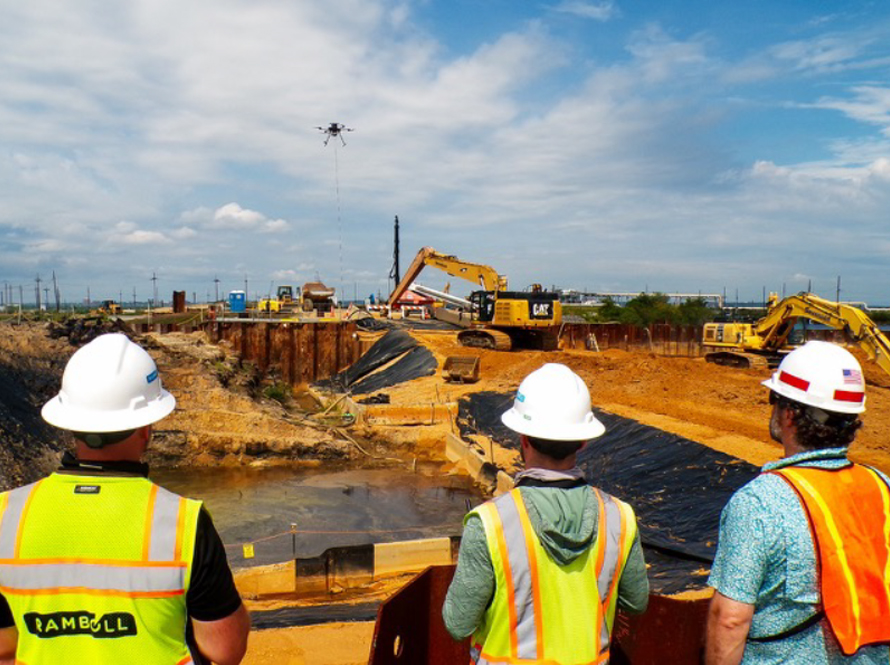 3 white males in safety gear watching a drone fly through the air.